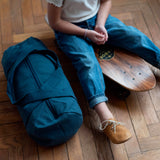 enfant assis sur un skateboard avec son sac de sport bleu pétrole posé sur sa droite